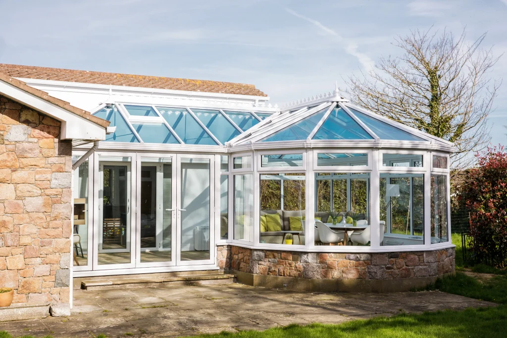 Exterior of a classic Victorian white UPVC conservatory with a red brick dwarf wall, decorative roof cresting, and arched window details, integrated into a landscaped garden with a stone patio and climbing roses.